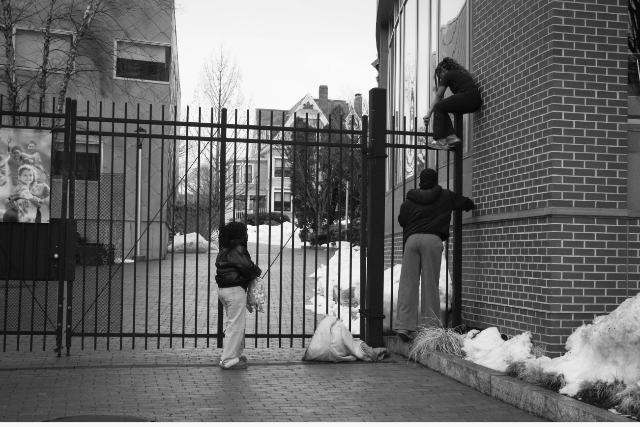 "Fencehopping" (2026), Canon EOS 5D Mark IV — a black-and-white documentary photograph of a child at a wrought-iron gate as two figures climb the fence beside a brick building in winter.