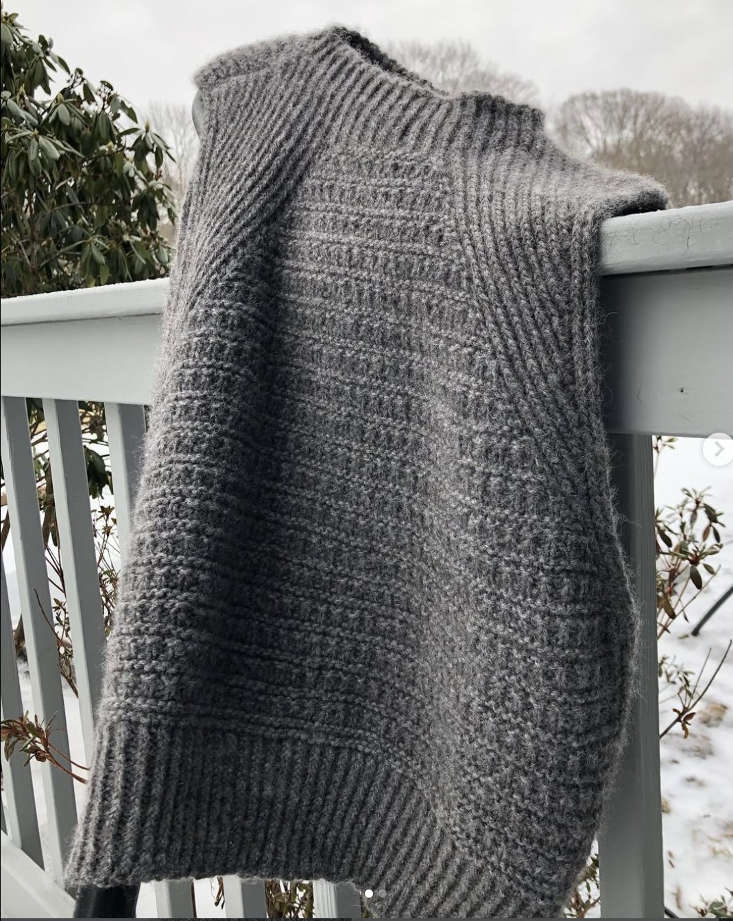 A grey knitted sweater vest draped over a white wooden railing outdoors in winter.