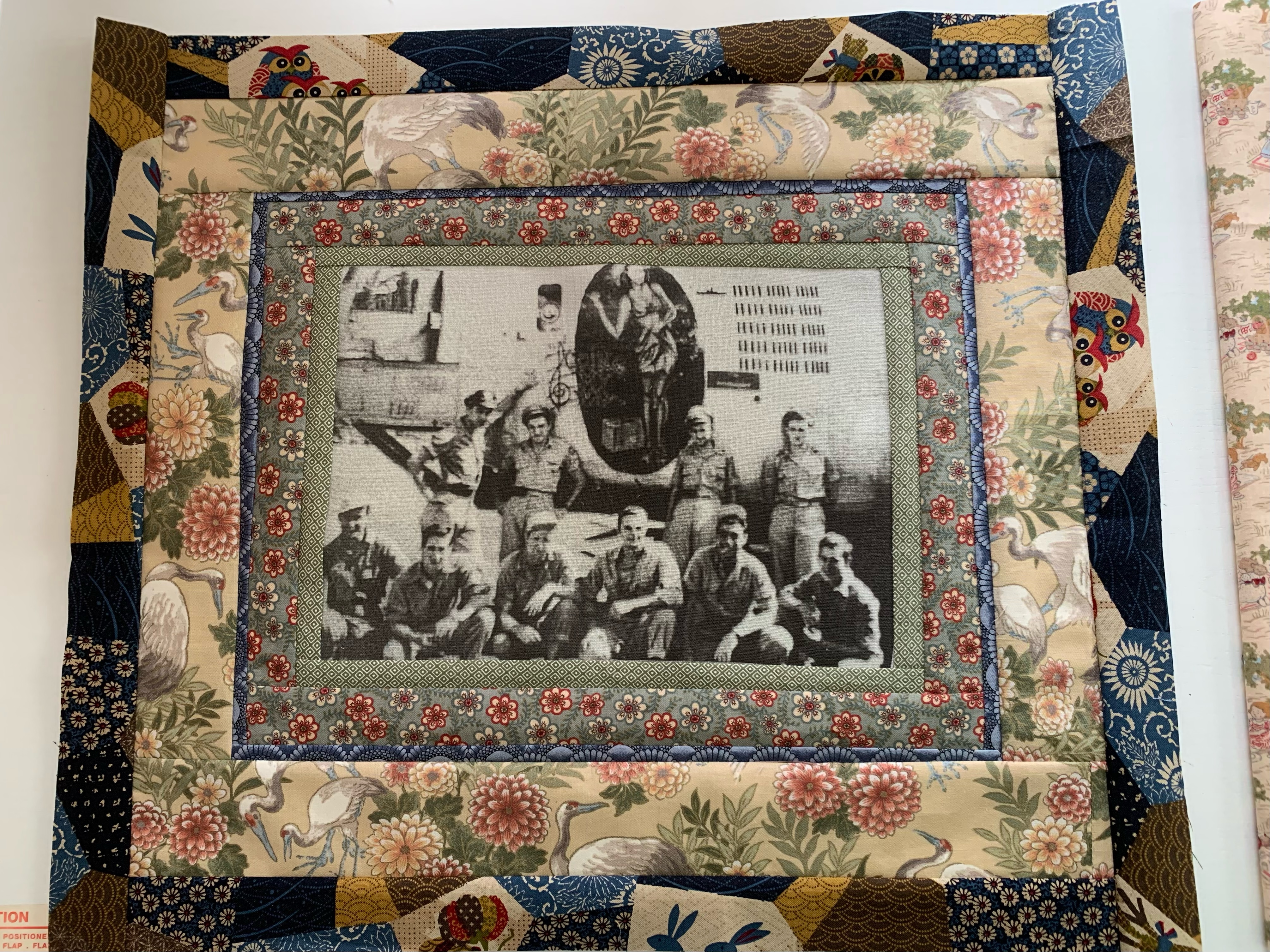 Quilted fabric wall hanging with a central black-and-white photograph of soldiers, framed by borders of floral, crane, and geometric patterned fabrics.