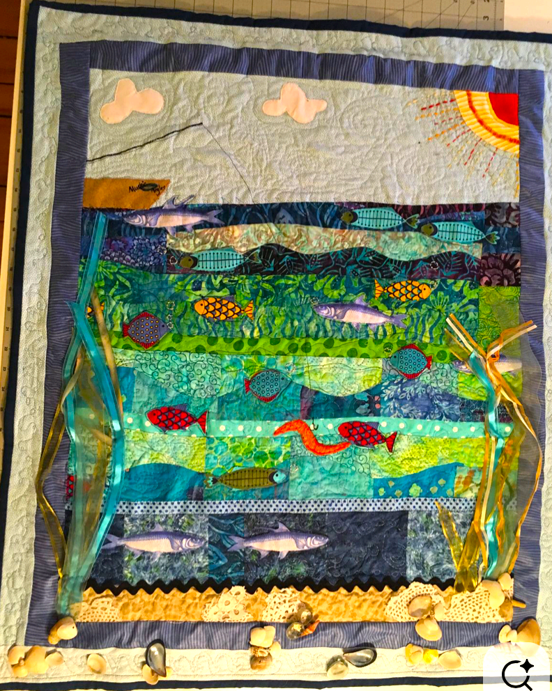 A quilted coastal scene with a sun, clouds, and layered rows of fish swimming through seaweed, trimmed with ribbons and small shells along the bottom.