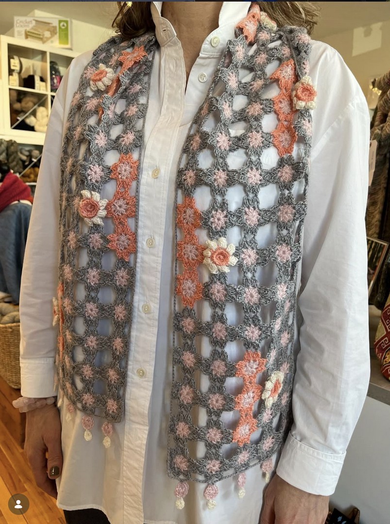 A person wearing a white button-down shirt with a long grey crocheted scarf featuring small pink and white flower accents draped over their shoulders.