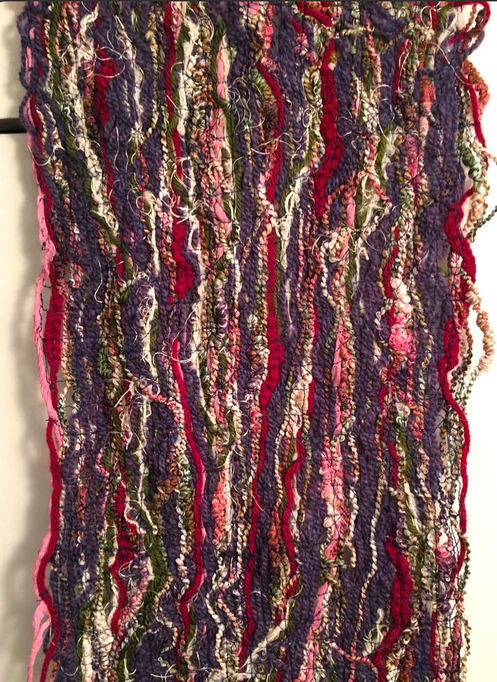 A textured hand-woven wall hanging combining purple, red, pink, green, and cream yarns of varied thicknesses and loops.