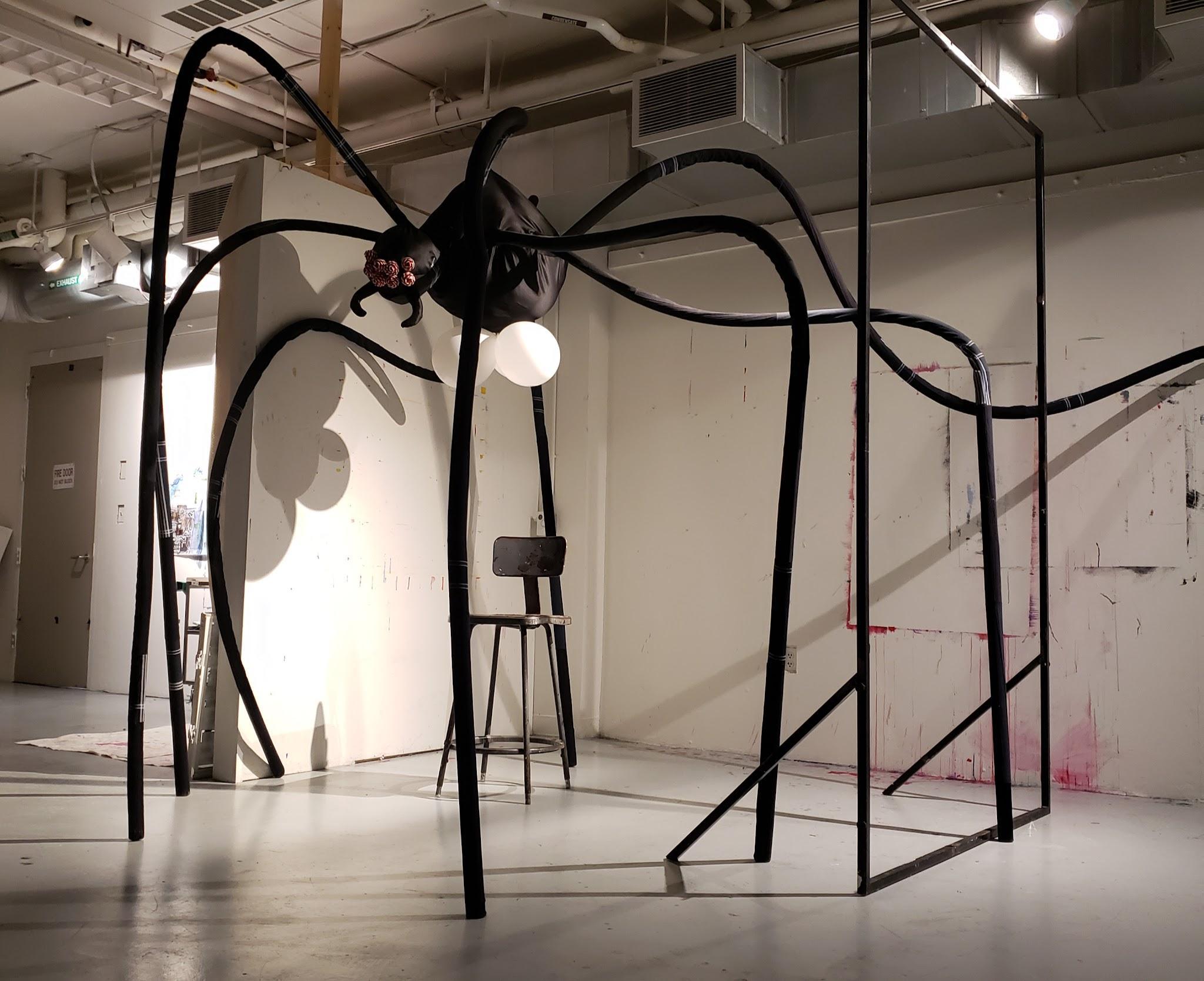 "Mom: After Louise Bourgeois", 2021; a large spider-like sculpture made of black fabric and pipe insulation standing in a gallery with a chair beneath it; ~10x10x10ft.