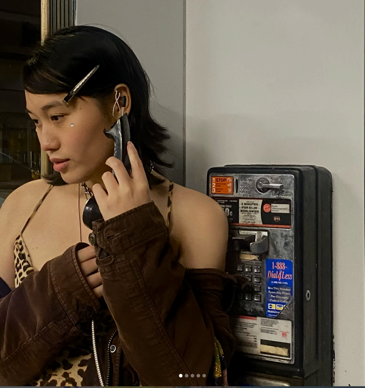 Self-portrait of the artist holding a payphone receiver, wearing a brown corduroy jacket over a leopard-print top.